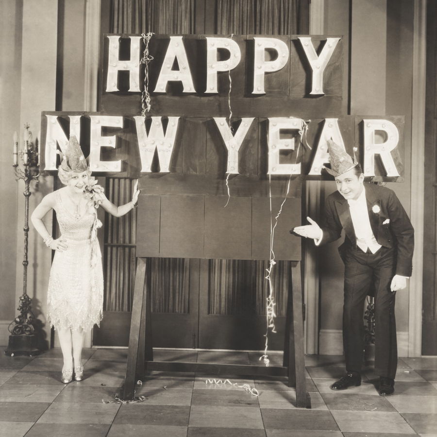 Eine alte schwarzweiß Aufnahme eines Paares am Silvesterabend vor einer Tafel auf der Happy New Year steht.