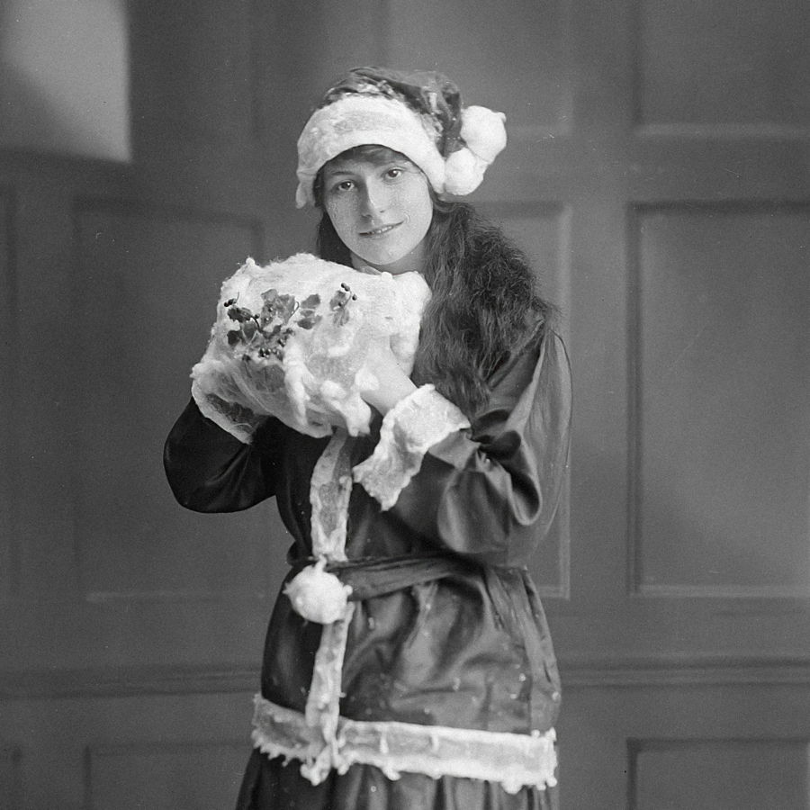 Eine alte schwarzweiß Aufnahme einer Frau, die als Weihnachtsmann verkleidet ist und ein Geschenk in der Hand hält.